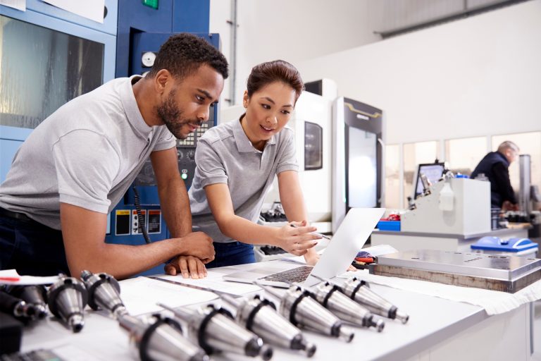 Two technicians in gray polo shirts review documents at a workbench with a laptop, surrounded by machining equipment in a factory.
