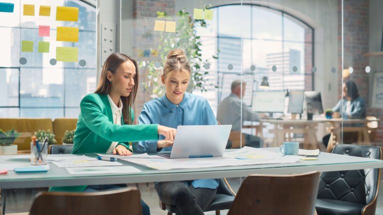 Two women collaborate at a bright office table, looking at a laptop with sticky notes on a glass wall behind them.