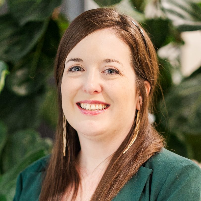 Smiling woman with long brown hair in a green blazer, indoor plants in the background (professional headshot).